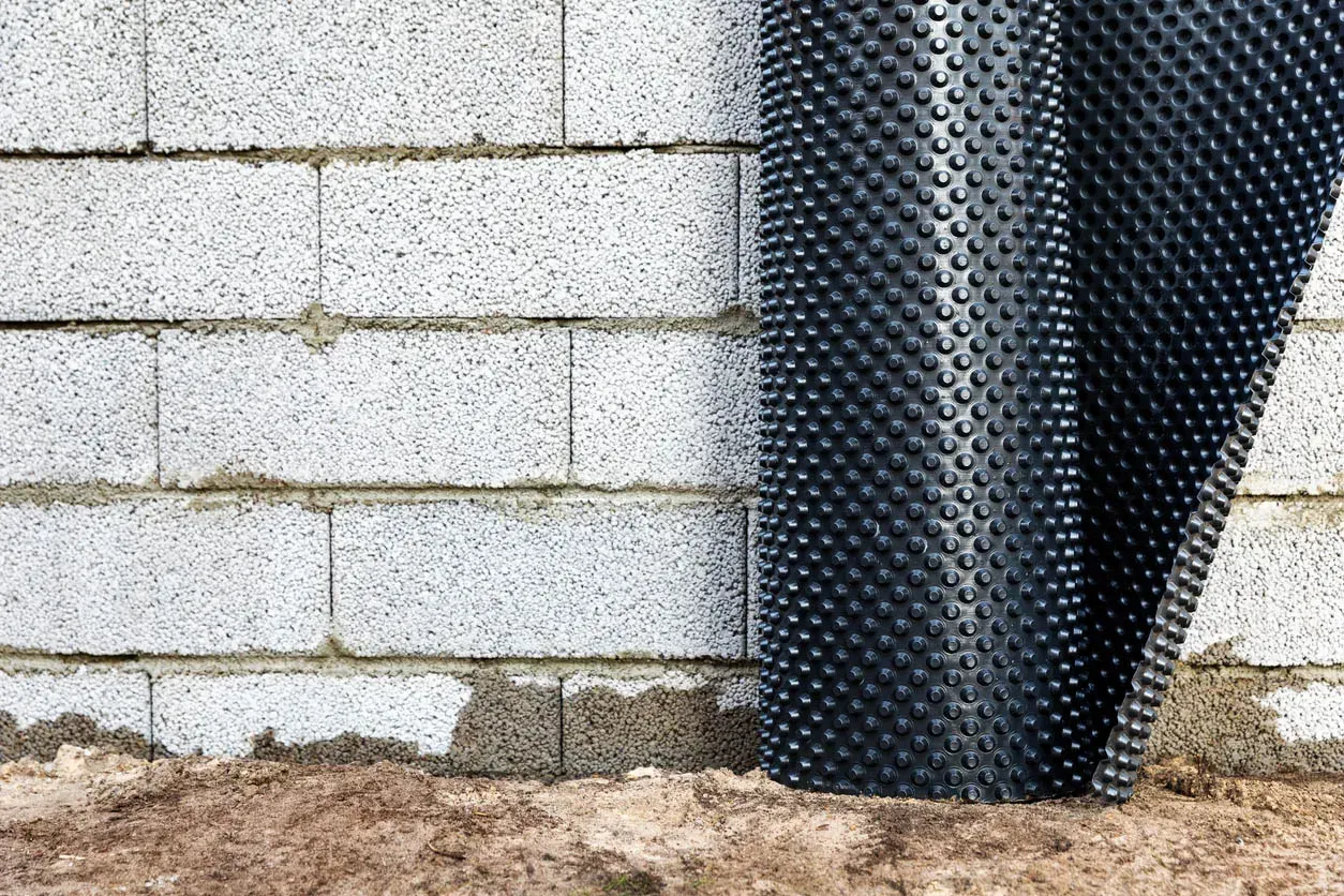 A roll of dimpled drainage membrane standing against a concrete block foundation wall